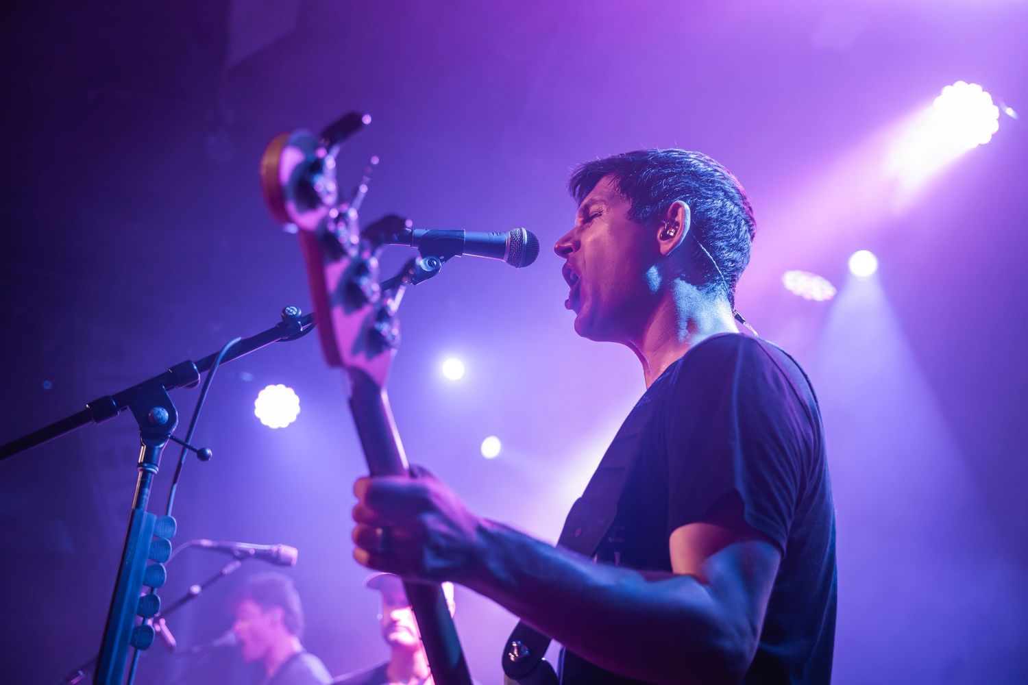 A musician performs on stage with a guitar and microphone against a purple-lit backdrop during a concert performance.