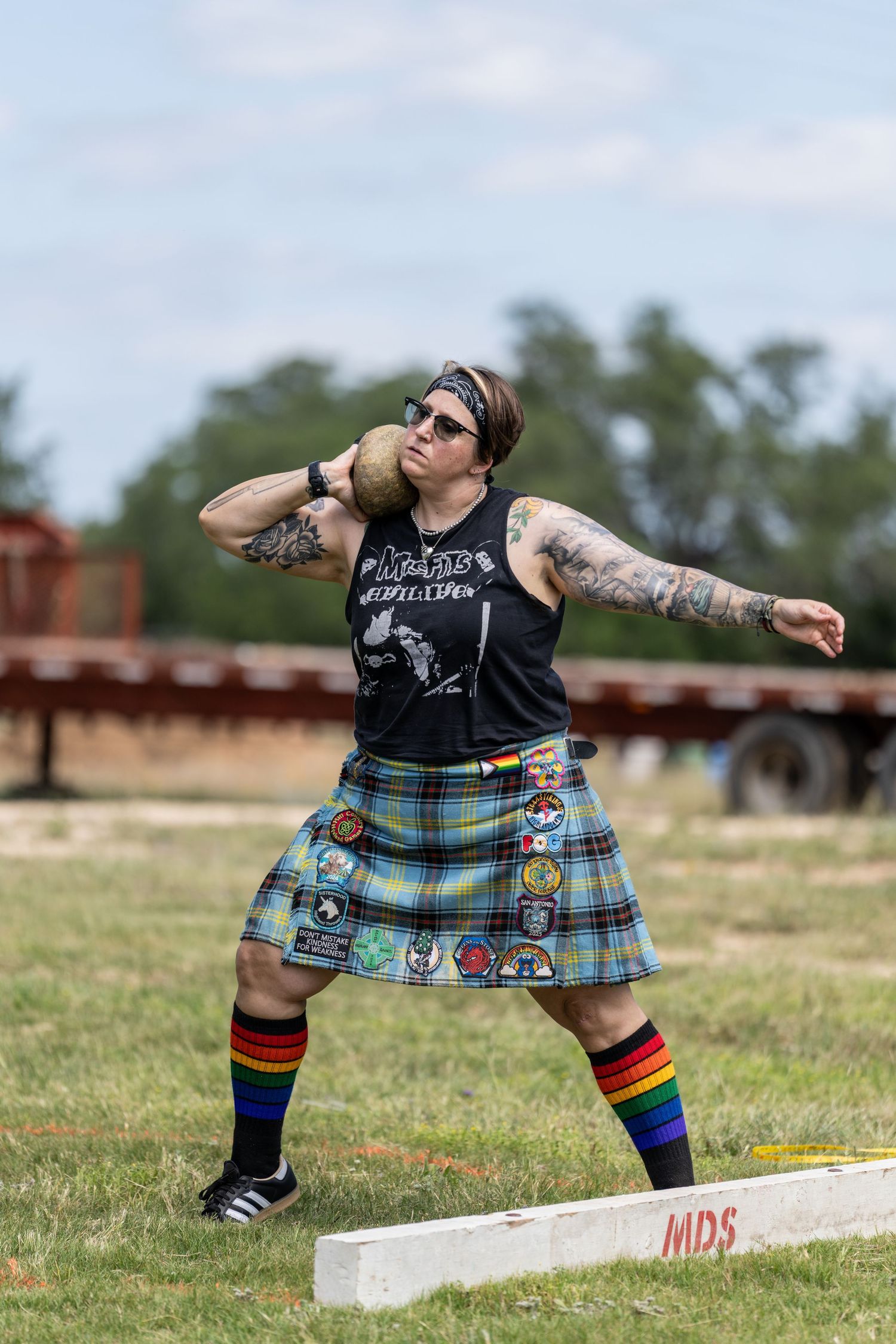 Tattooed athlete in plaid kilt throws shot put at Highland Games competition outdoors.
