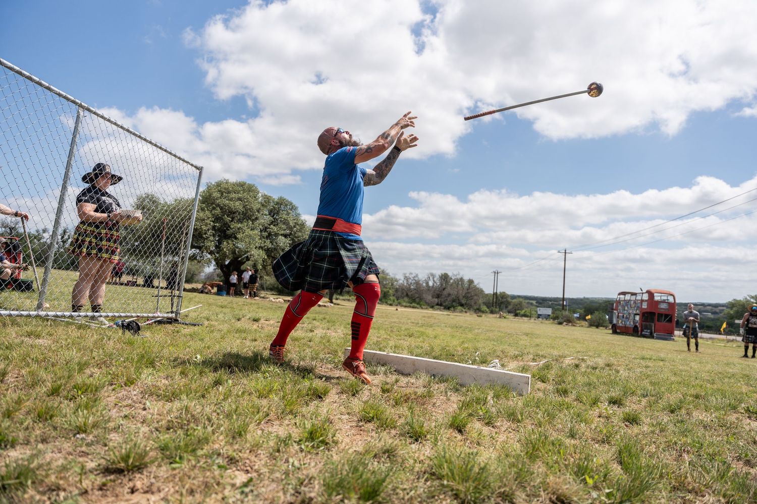 Man in kilt and red socks throws hammer at Highland Games competition outdoors on sunny day.