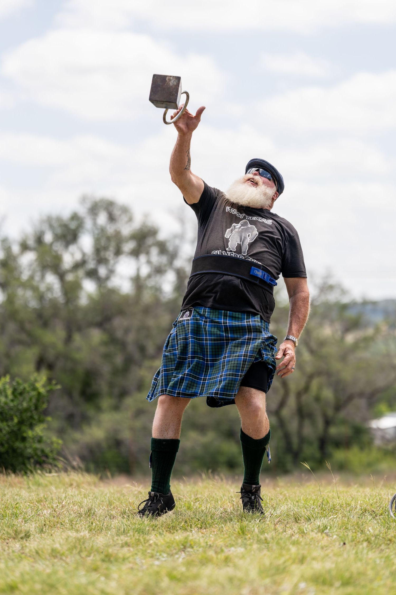 Bearded man in blue tartan kilt throwing weight at Scottish Highland Games competition outdoors.