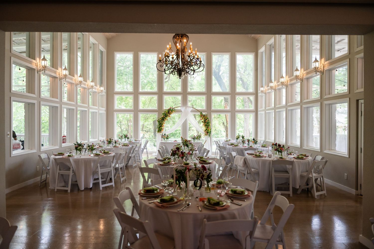 Elegant wedding reception hall with white round tables, floral centerpieces, chandelier, and large windows overlooking greenery.