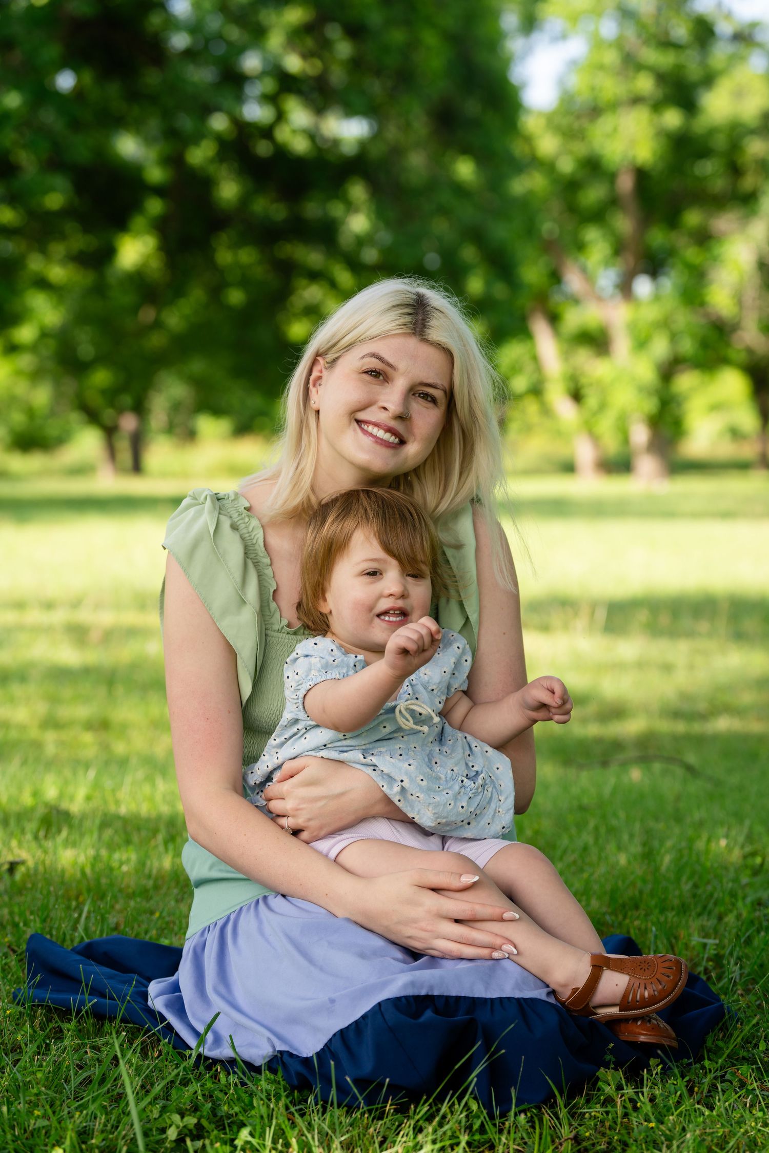 Smiling blonde mother sitting on grass in park holding toddler daughter in floral dress on sunny day.