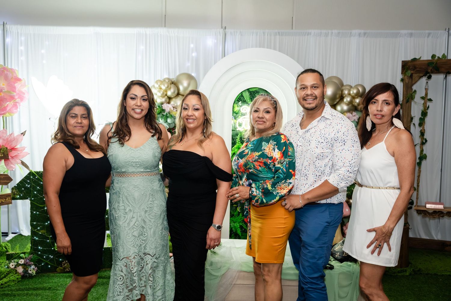 Group of six adults posing together in front of a floral arch backdrop at a quinceañera celebration.
