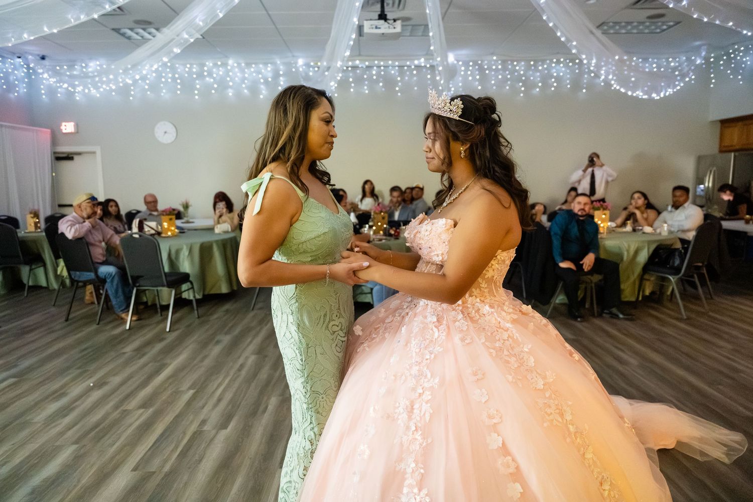 Two women share a dance at a quinceañera celebration in a decorated banquet hall with guests seated at tables.