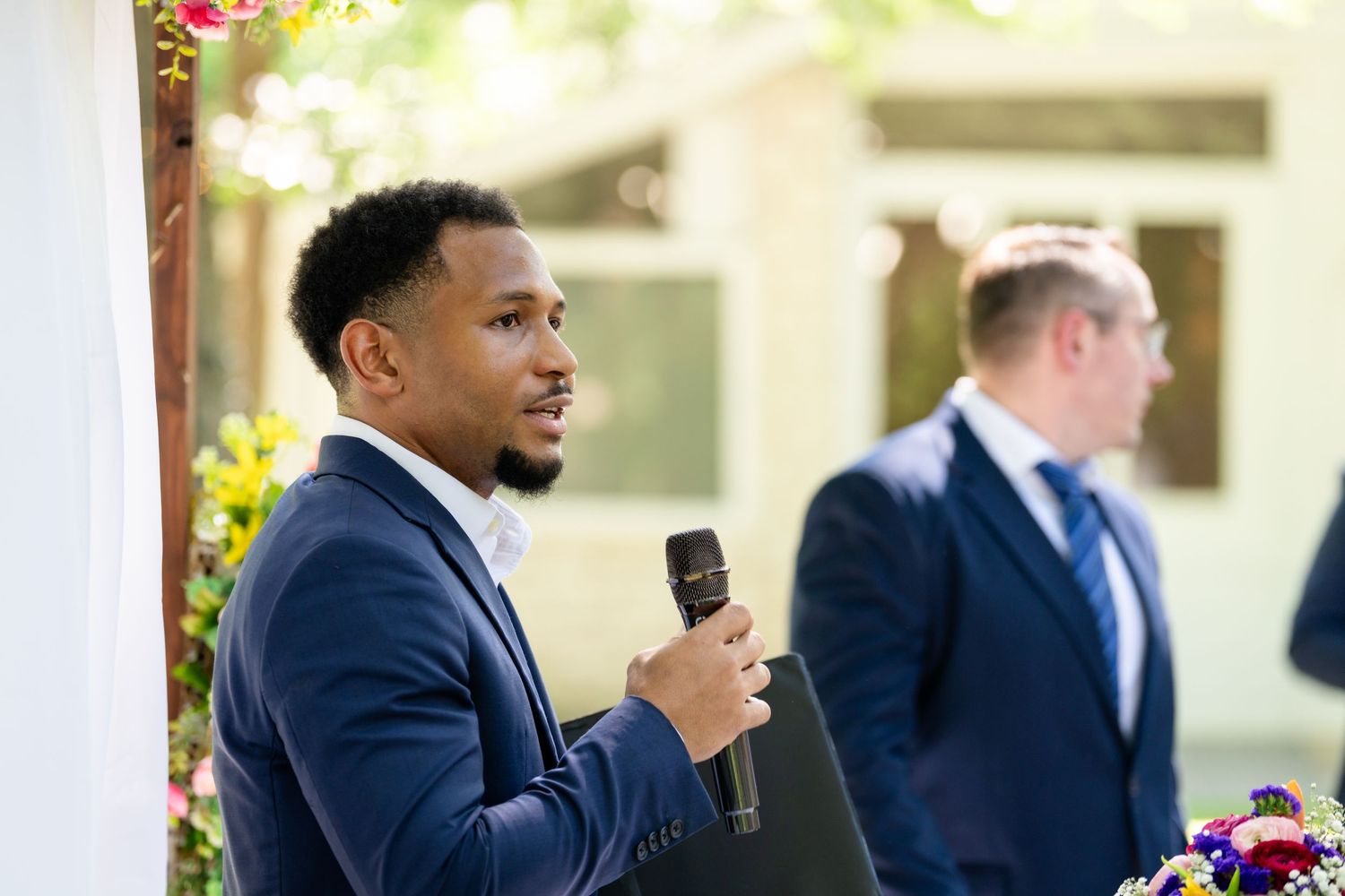 Man in navy suit speaking into microphone at outdoor wedding ceremony with guest in background.