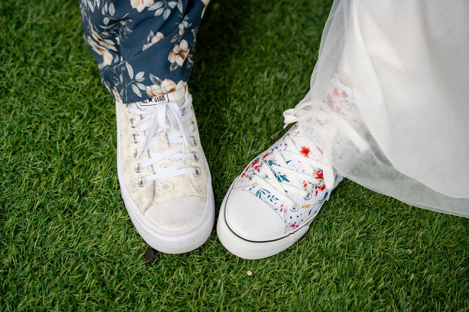 Close-up of two people wearing white sneakers on green grass, one plain and one with colorful floral designs, beside a white wedding dress.