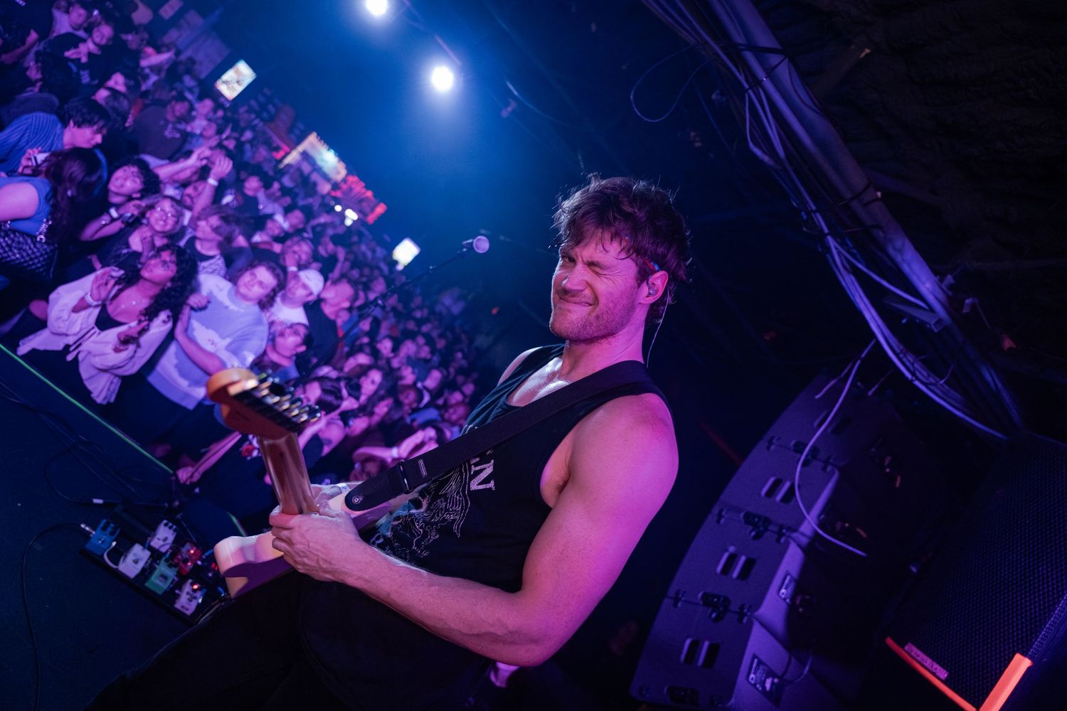 Muscular guitarist performing on stage under purple lights with an energetic crowd behind him at a live concert.