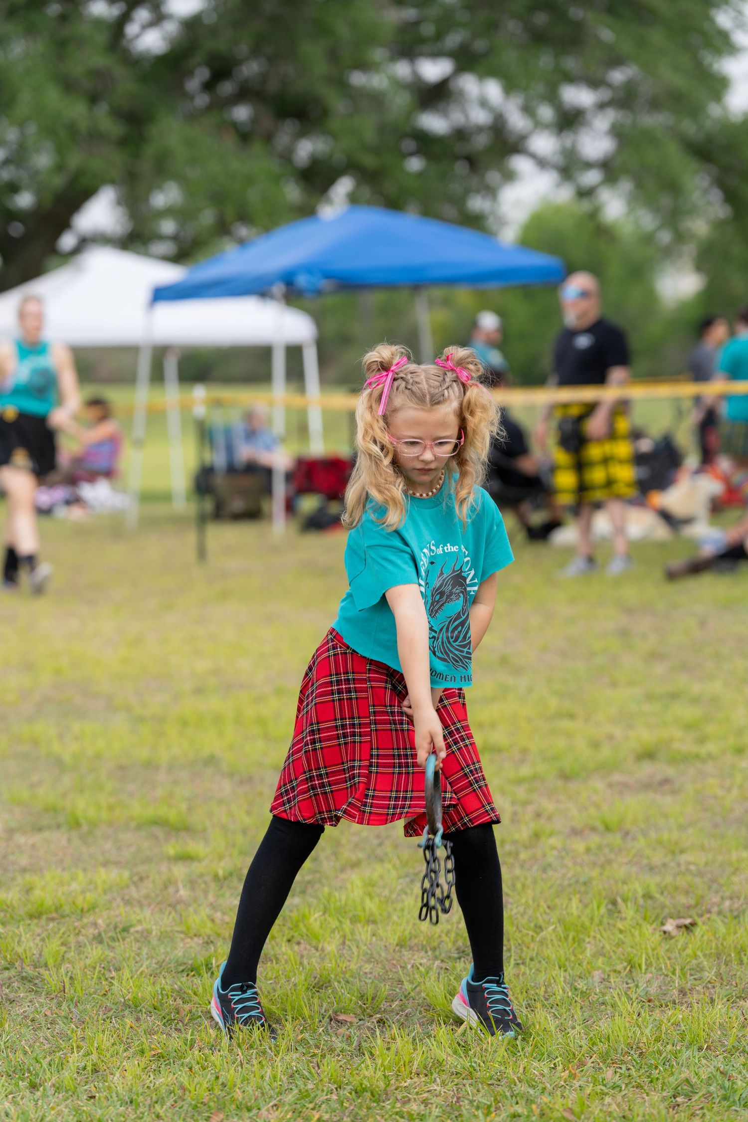 Young girl in red tartan skirt and teal shirt performing Scottish Highland dance on grass at outdoor festival.