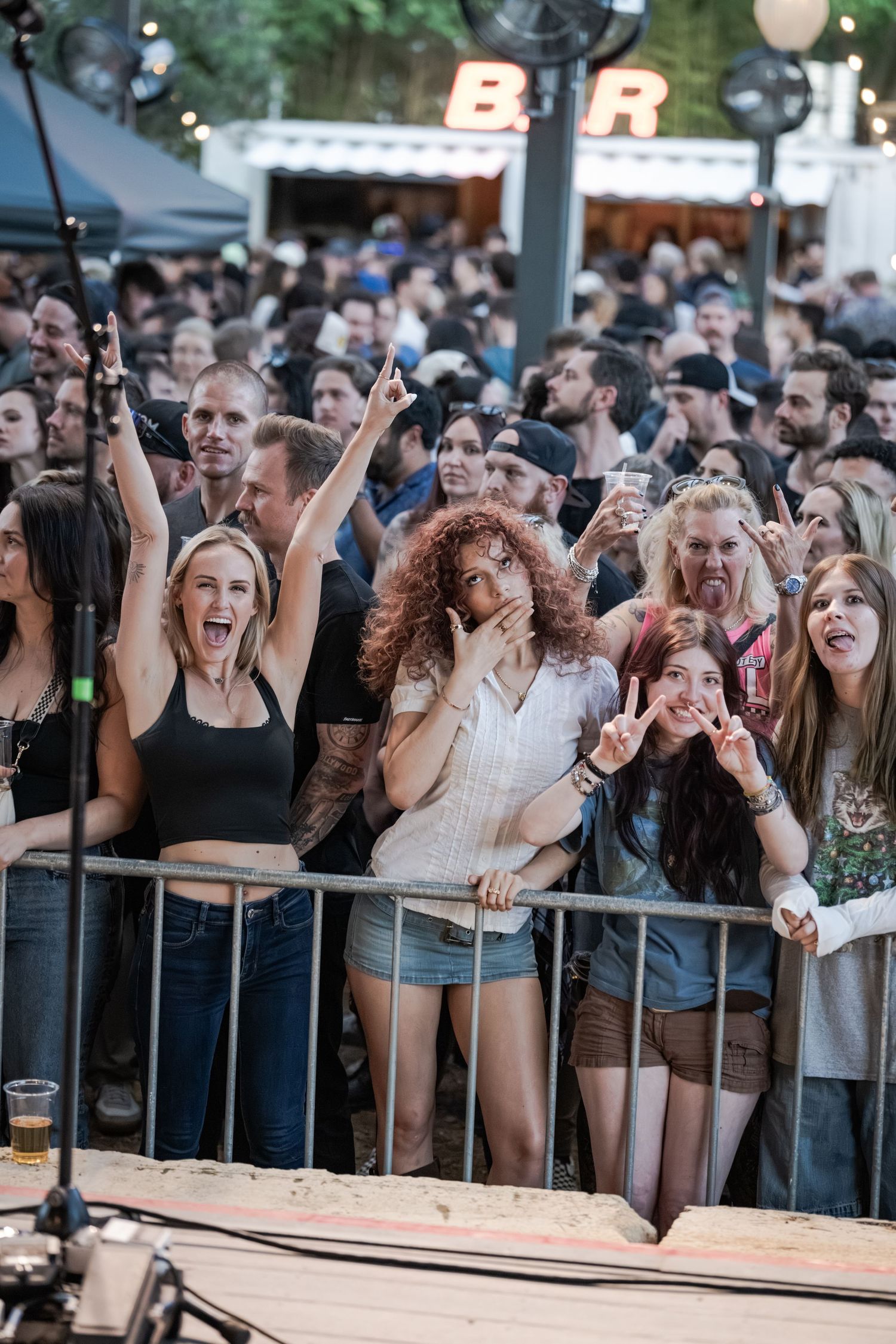 Excited crowd of fans at an outdoor concert, with women cheering and making rock signs at the stage barrier.