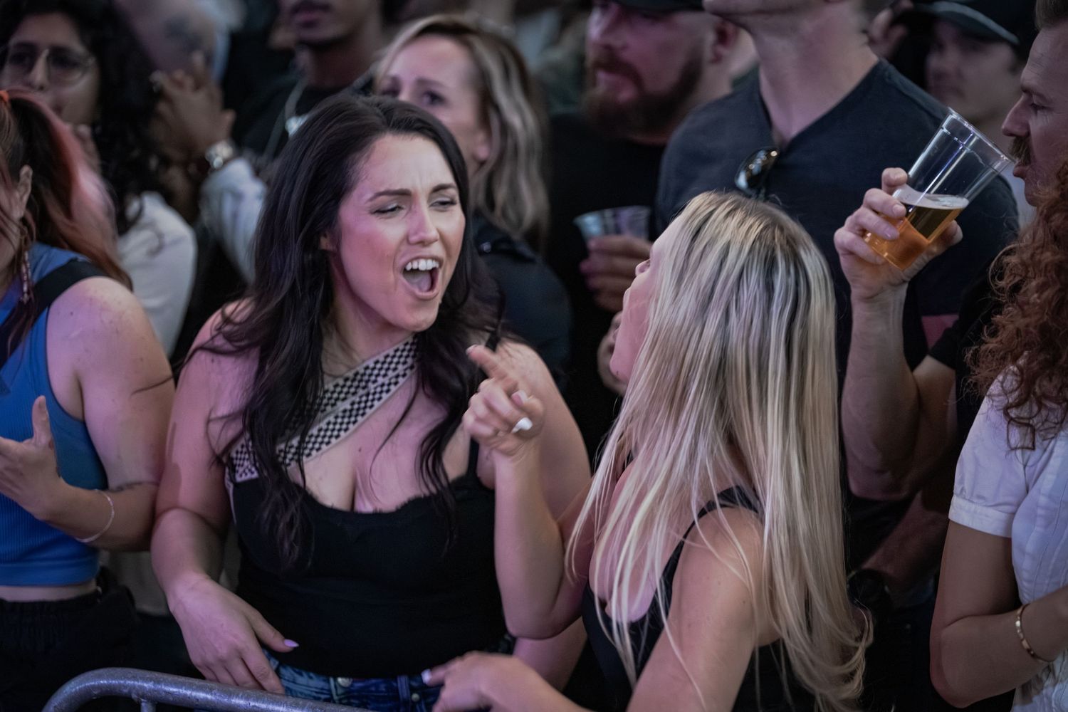 Two women laughing and enjoying drinks at an outdoor concert event crowd.