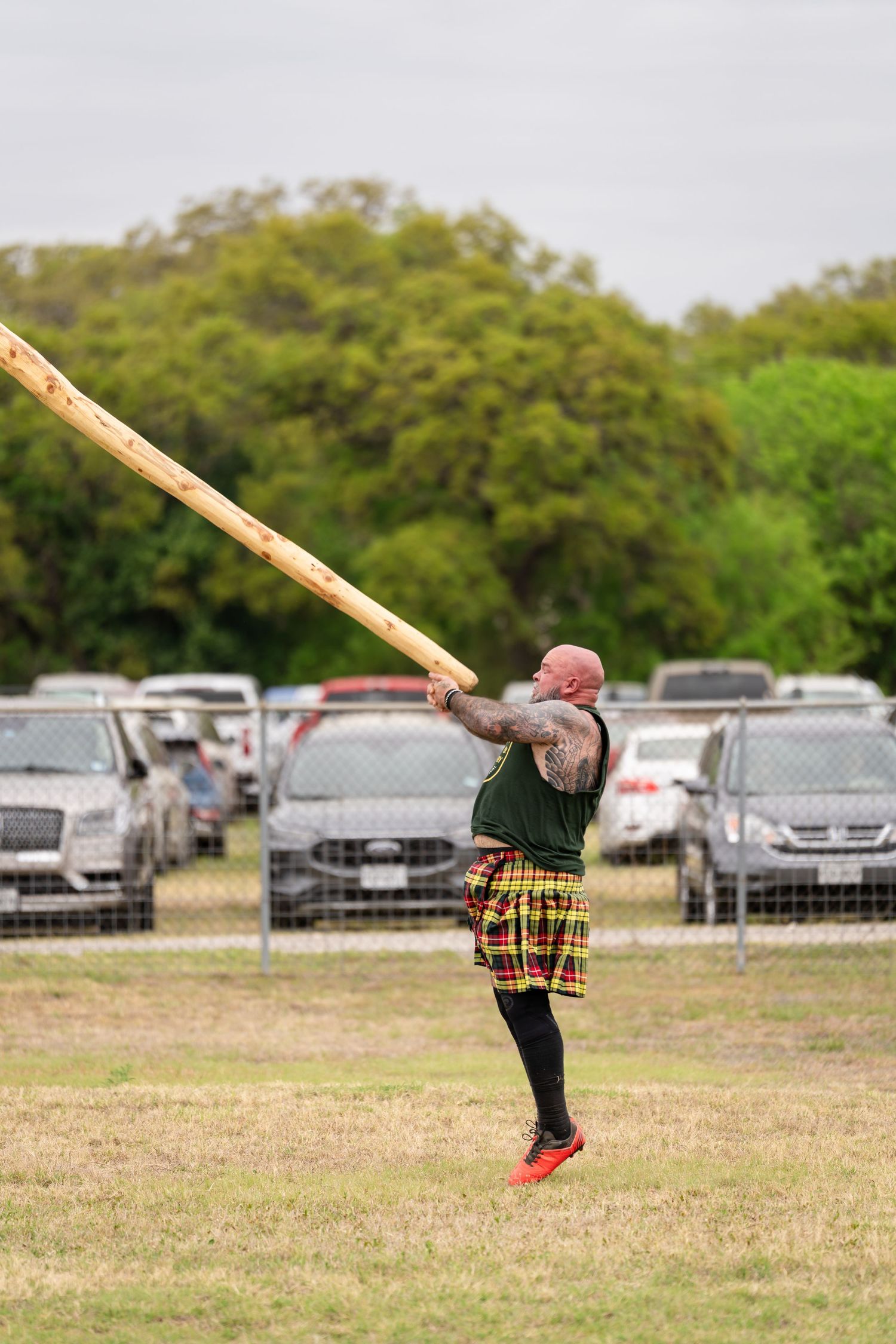 Tattooed athlete in tartan kilt tossing a caber at a Highland Games competition outdoors.