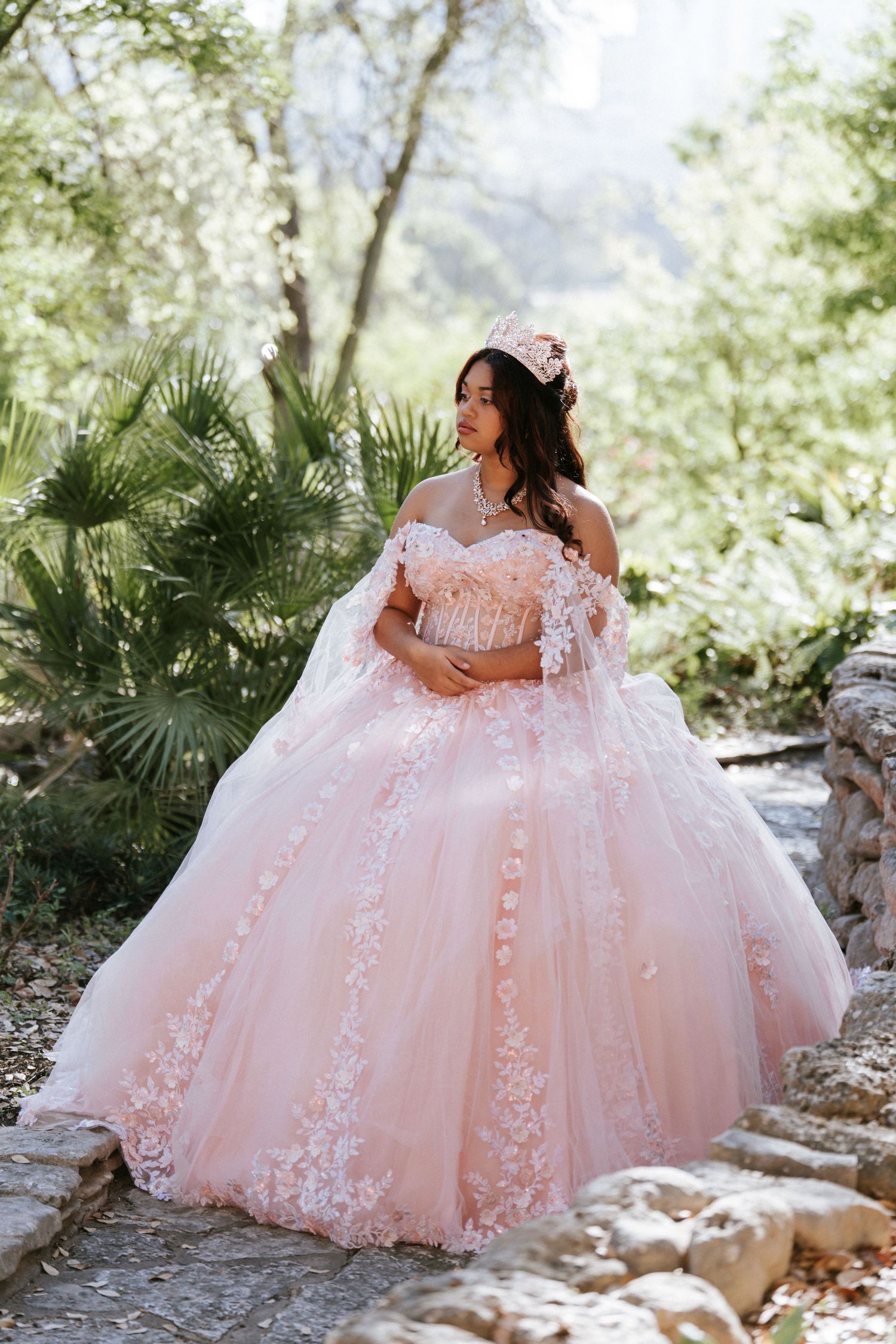 Young woman in a blush pink ball gown with lace appliques and floral crown poses outdoors for quinceañera photos.
