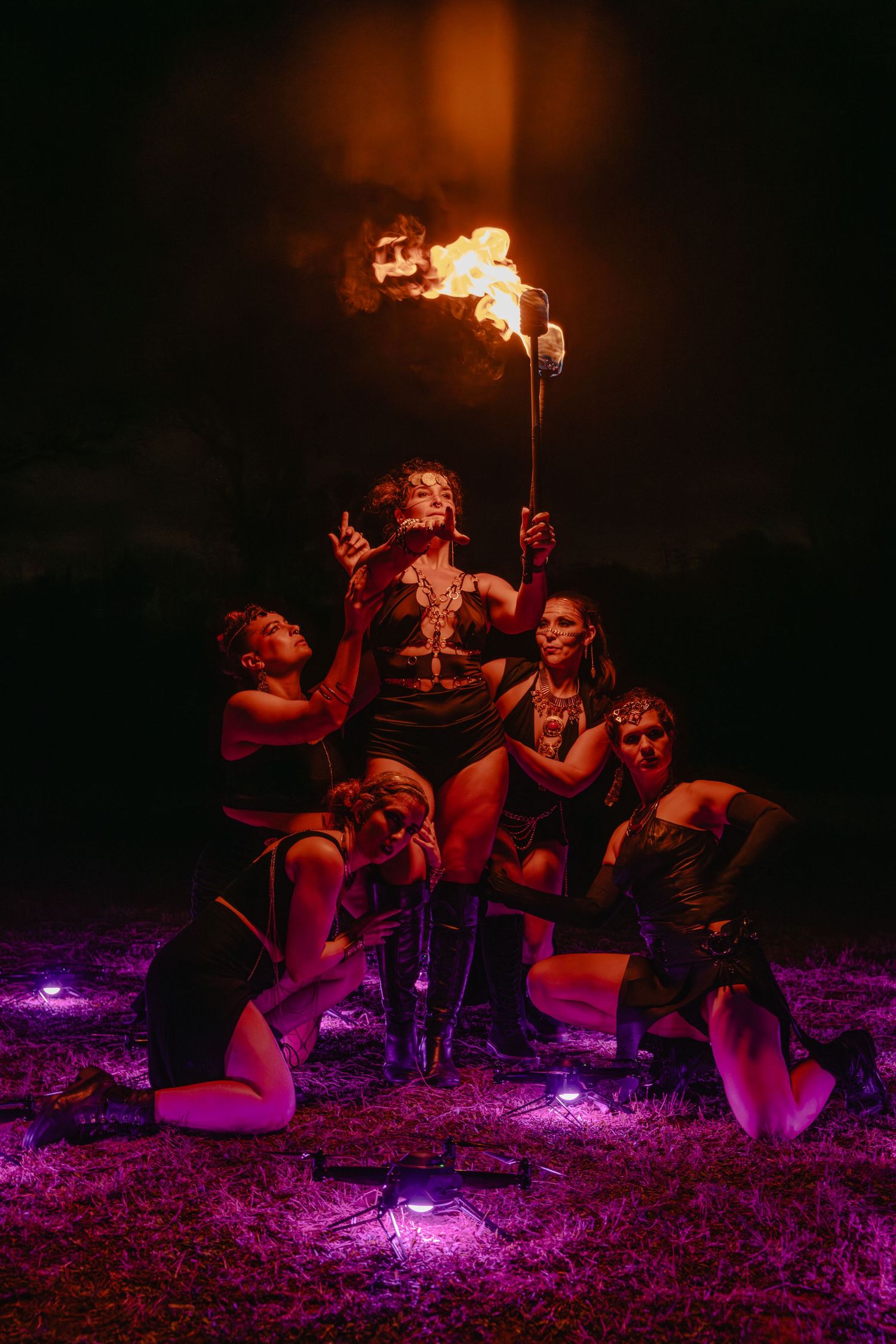Fire performers pose dramatically at night, with one holding a flaming torch, surrounded by purple lights on grass.