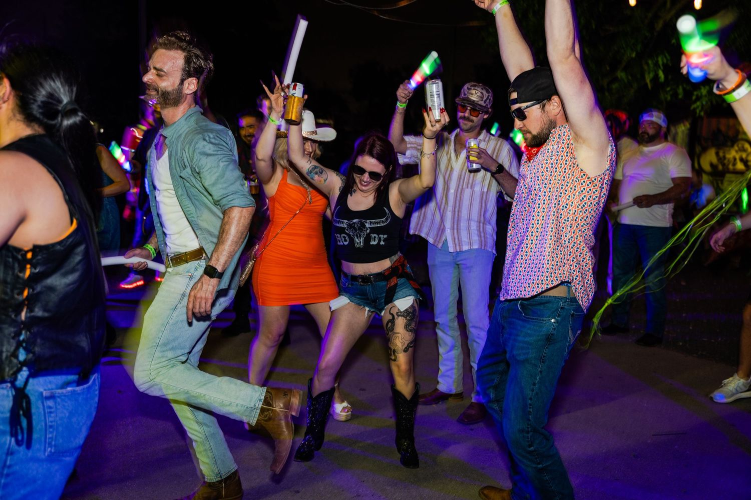 Group of friends dancing and celebrating at an outdoor party with colorful glow sticks at night.