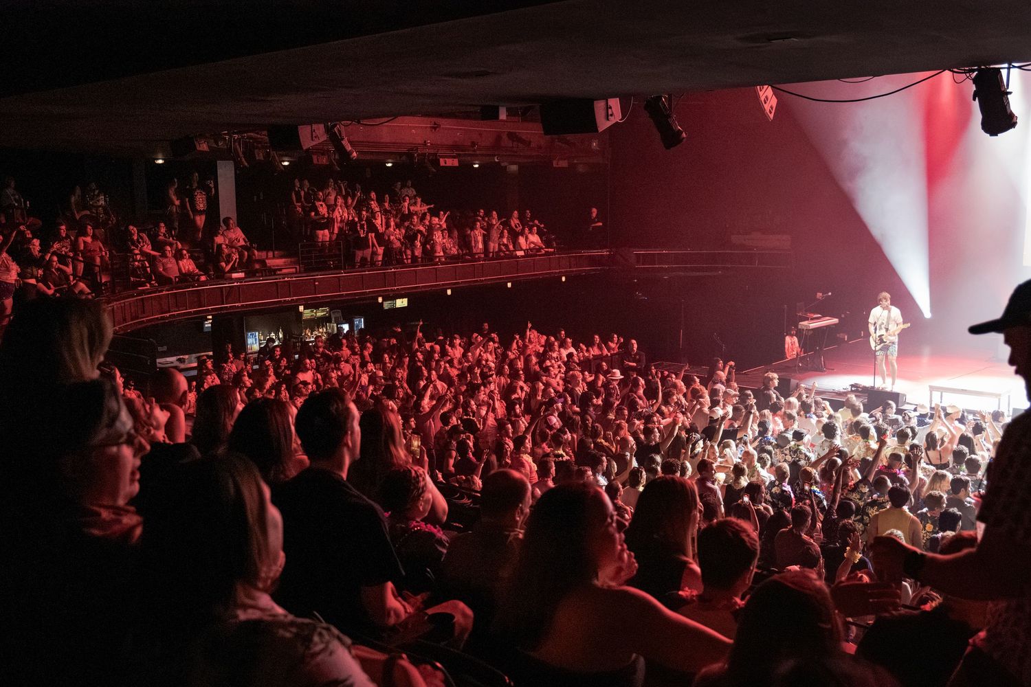 A packed concert venue bathed in red light with a large crowd facing a performer on a bright stage.