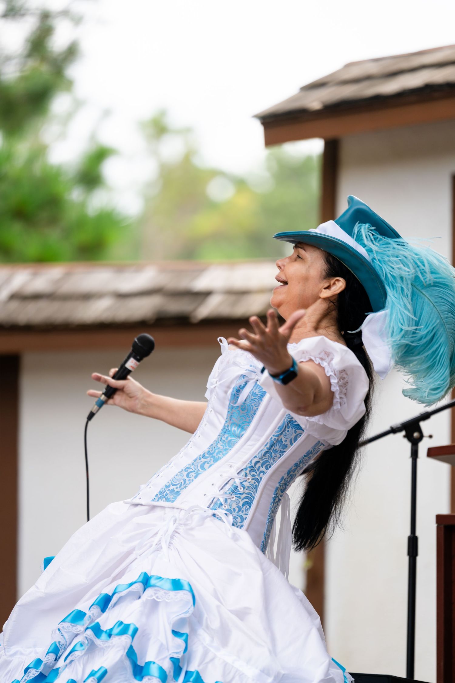 Woman in white and blue Victorian dress and teal feathered hat singing passionately on outdoor stage with microphone.