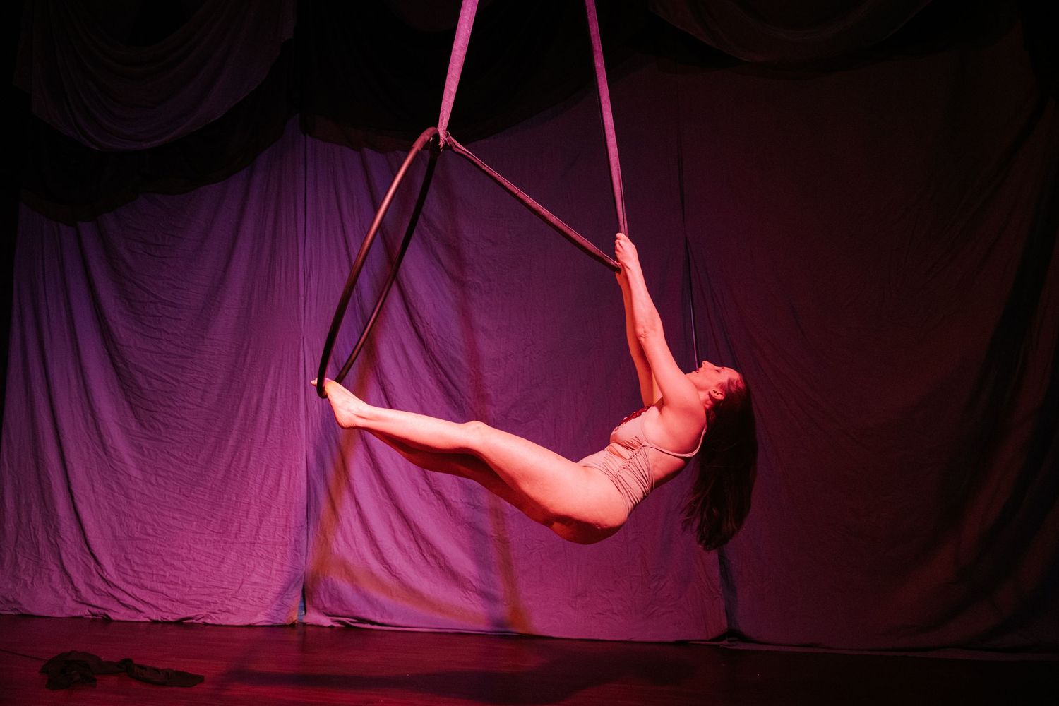 Aerial acrobat performing on pink silk hoop apparatus on stage under dramatic purple and red lighting.