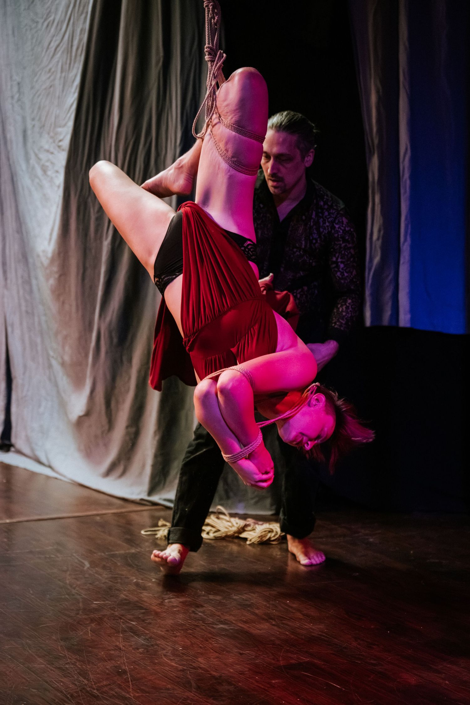 Aerial acrobat wrapped in red silk suspended upside down while male performer supports her on stage.