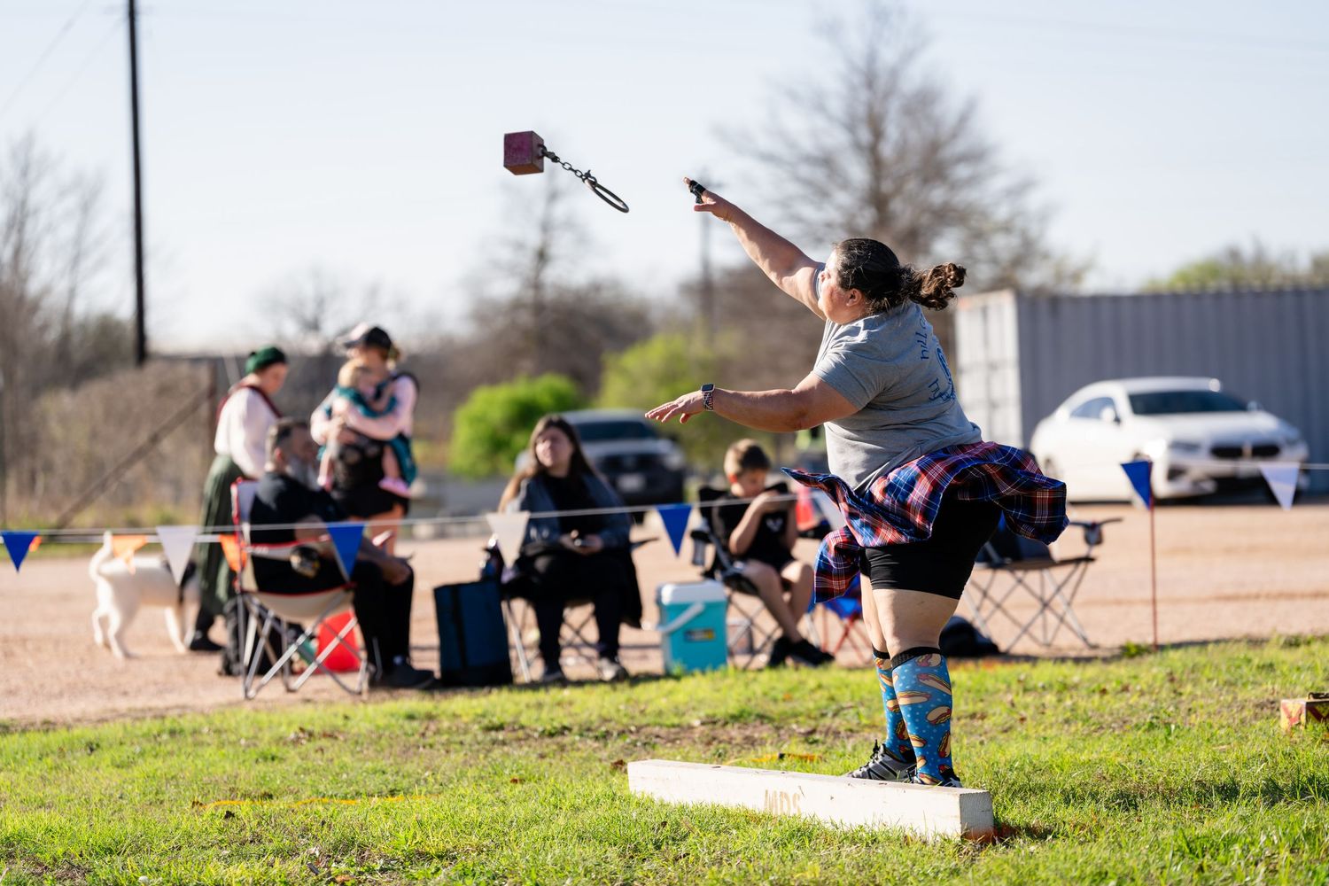 Woman throwing a weight in a Highland Games competition outdoors, with spectators seated in chairs watching nearby.