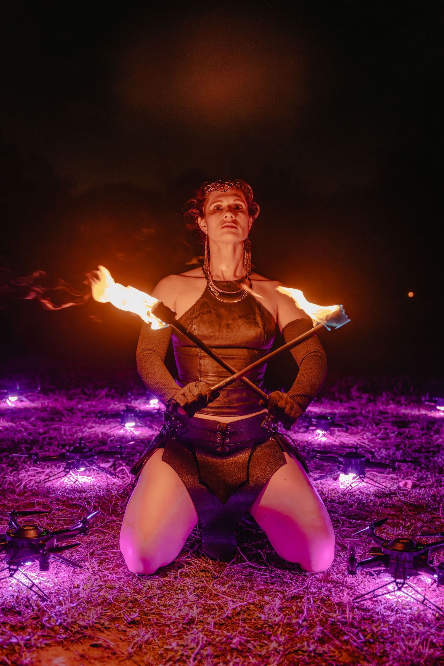 Fire performer kneeling on ground at night holding flaming batons surrounded by purple lights during outdoor show.