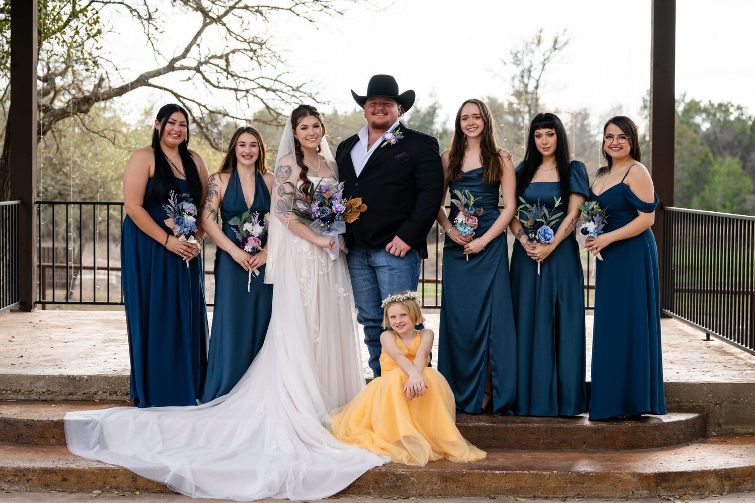 Wedding party poses outdoors, bride in white gown, groom in black hat, bridesmaids in teal dresses, flower girl in yellow.