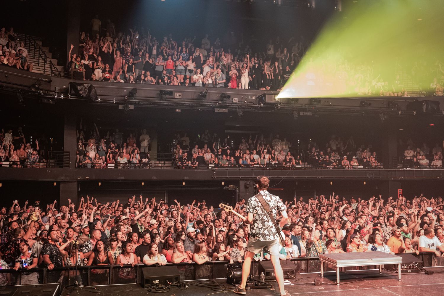 Performer on stage facing a large enthusiastic crowd in a packed concert theater venue.