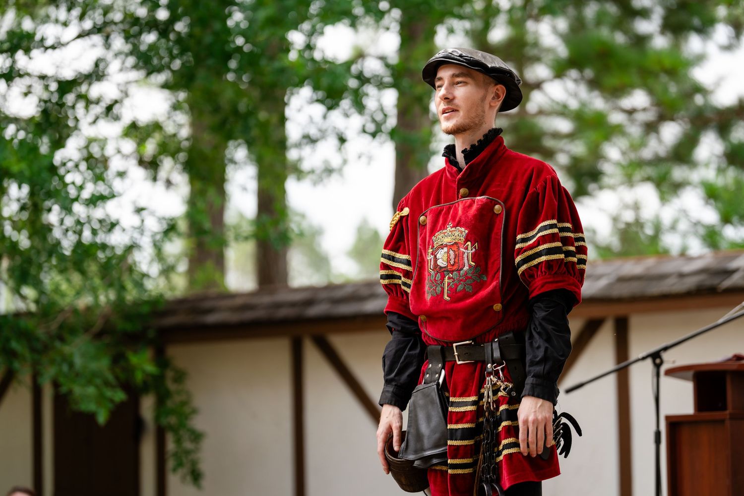 Man in red medieval costume with emblem stands outdoors near trees and building.