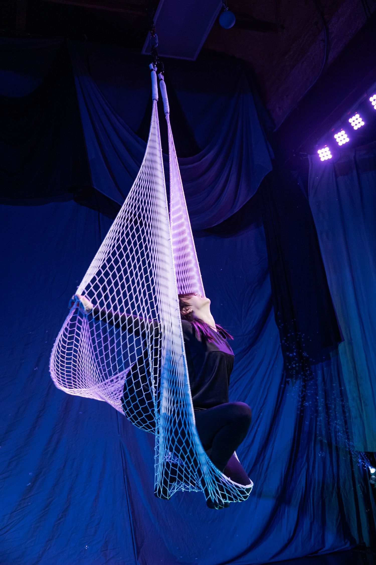 Aerial performer suspended in a white net hammock under blue stage lighting during a circus performance.