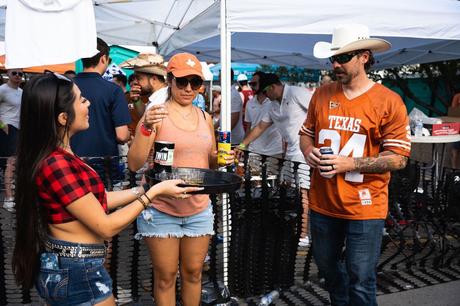 Tailgaters in sports gear socialize under white tents with drinks.