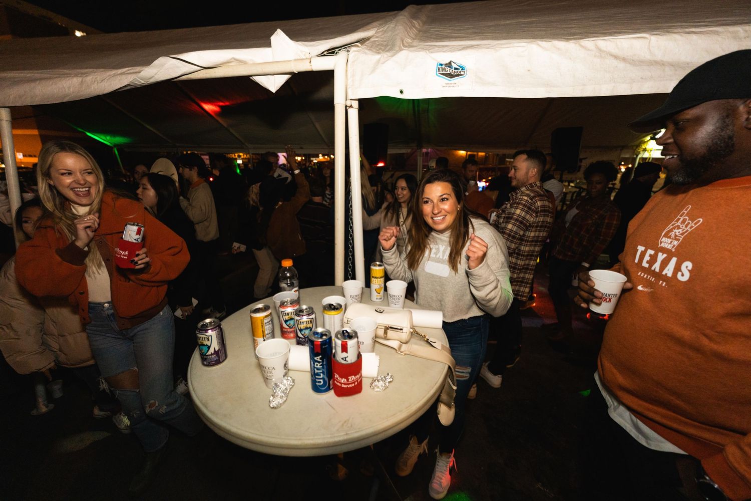 People gathered around table under tent at outdoor night event with drinks.