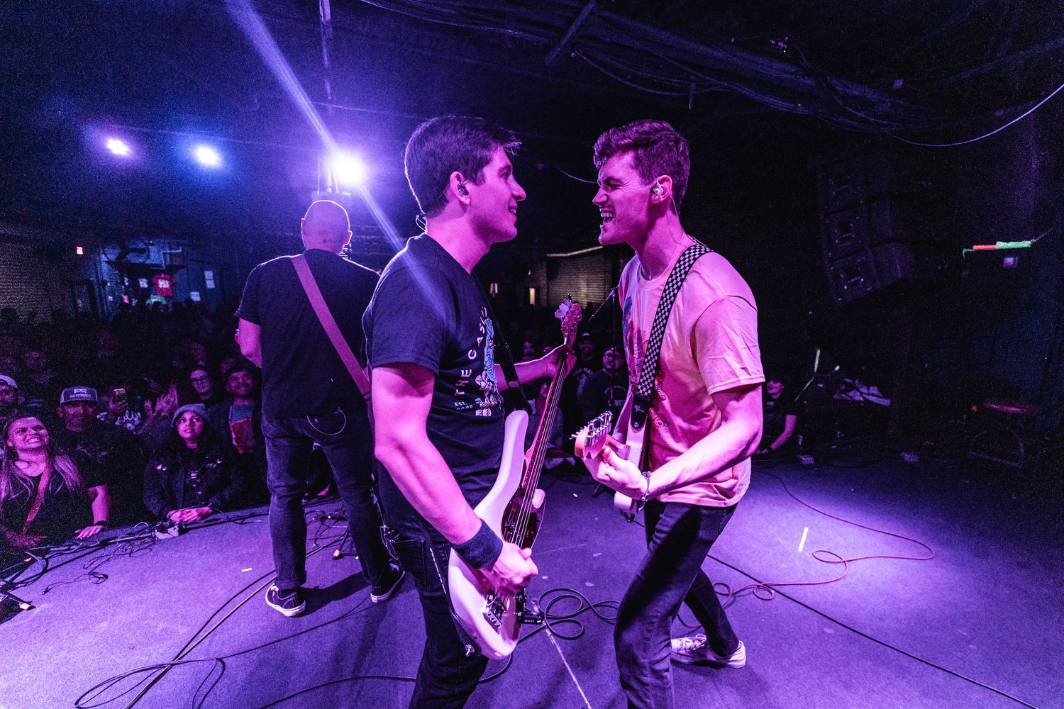 Musicians perform energetically on stage with purple lighting and crowd visible.
