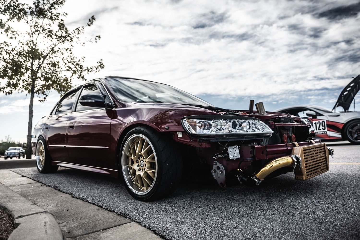 A maroon Subaru WRX STI with gold wheels parked at a car show under cloudy skies.