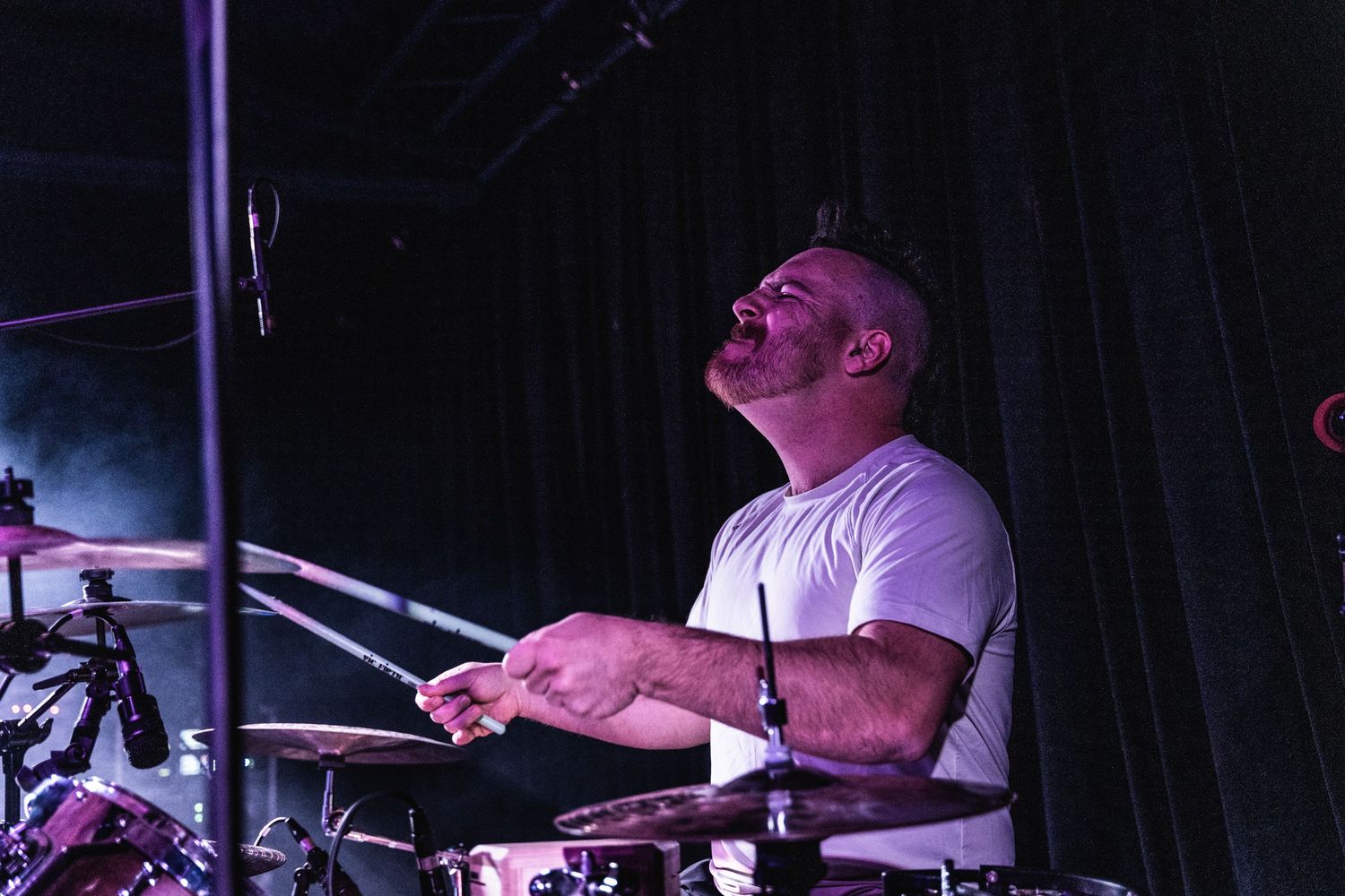 Drummer performing on stage under dramatic purple lighting at a concert venue.