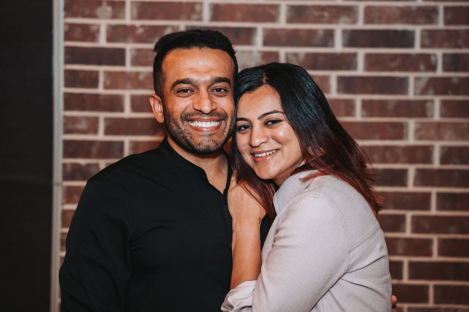 A couple embraces while smiling warmly against a brick wall background.
