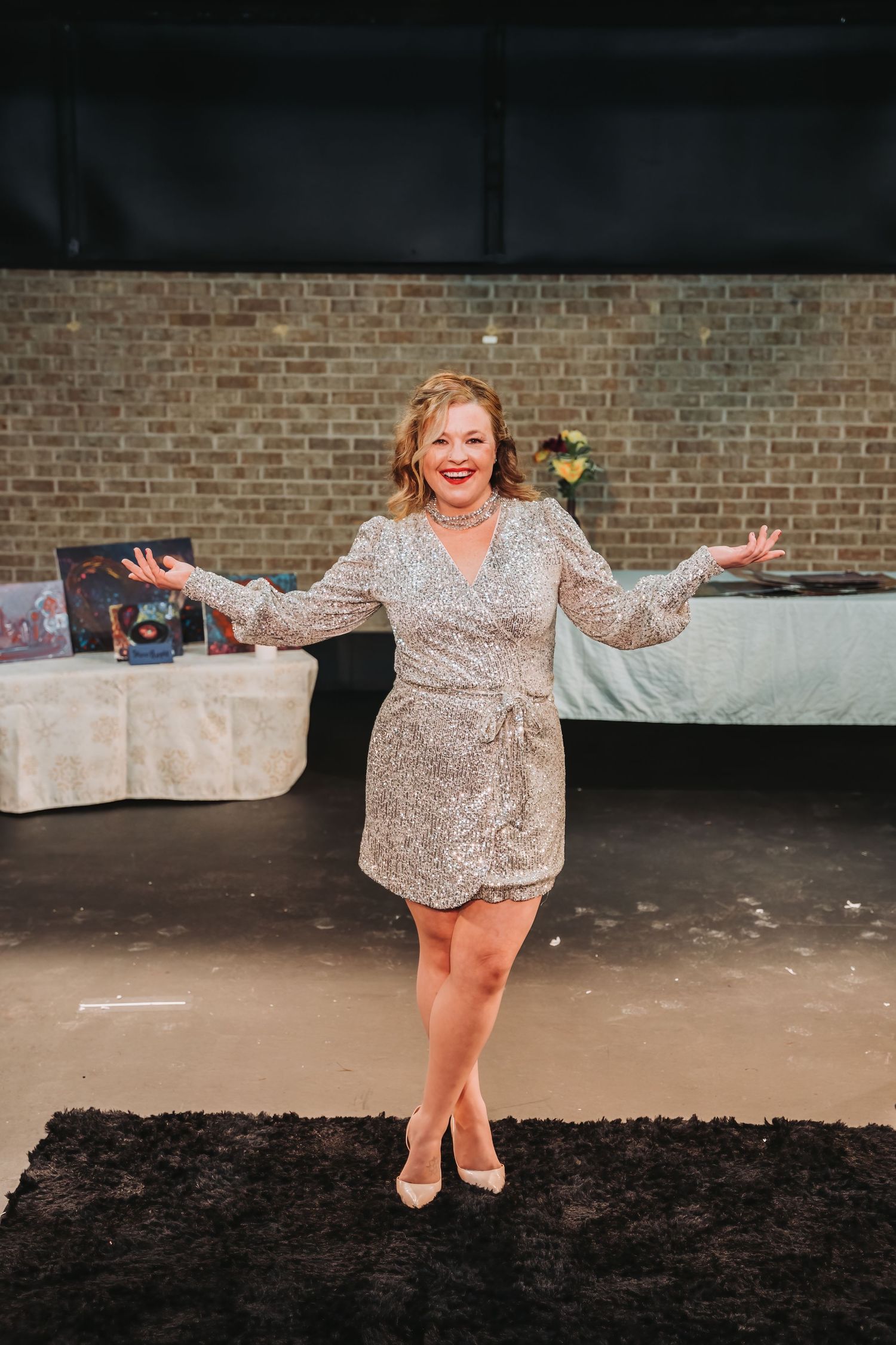 A performer in a sparkly silver dress poses joyfully on stage against a brick wall background.