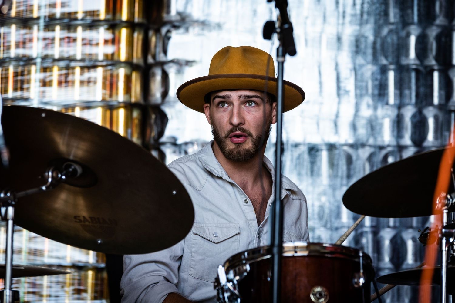 Drummer in a wide-brimmed hat performing behind a drum set in an industrial setting.