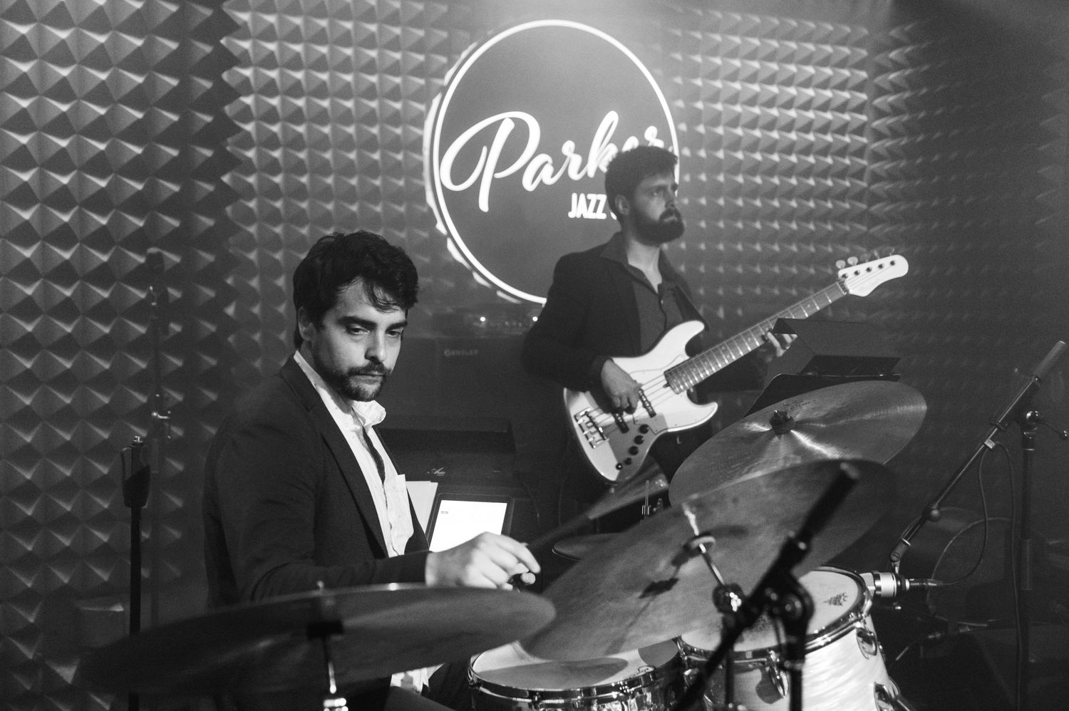 Black and white photo of musicians performing at Parker Jazz Club with drum kit and guitar visible.