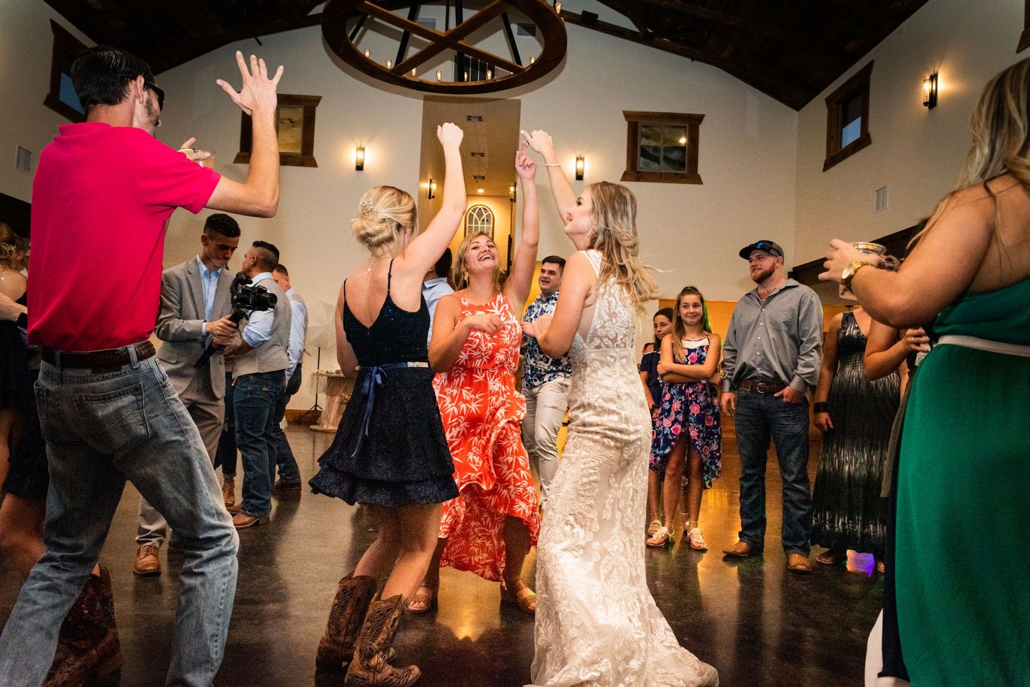 Wedding party dances enthusiastically during reception in rustic venue with high ceilings.