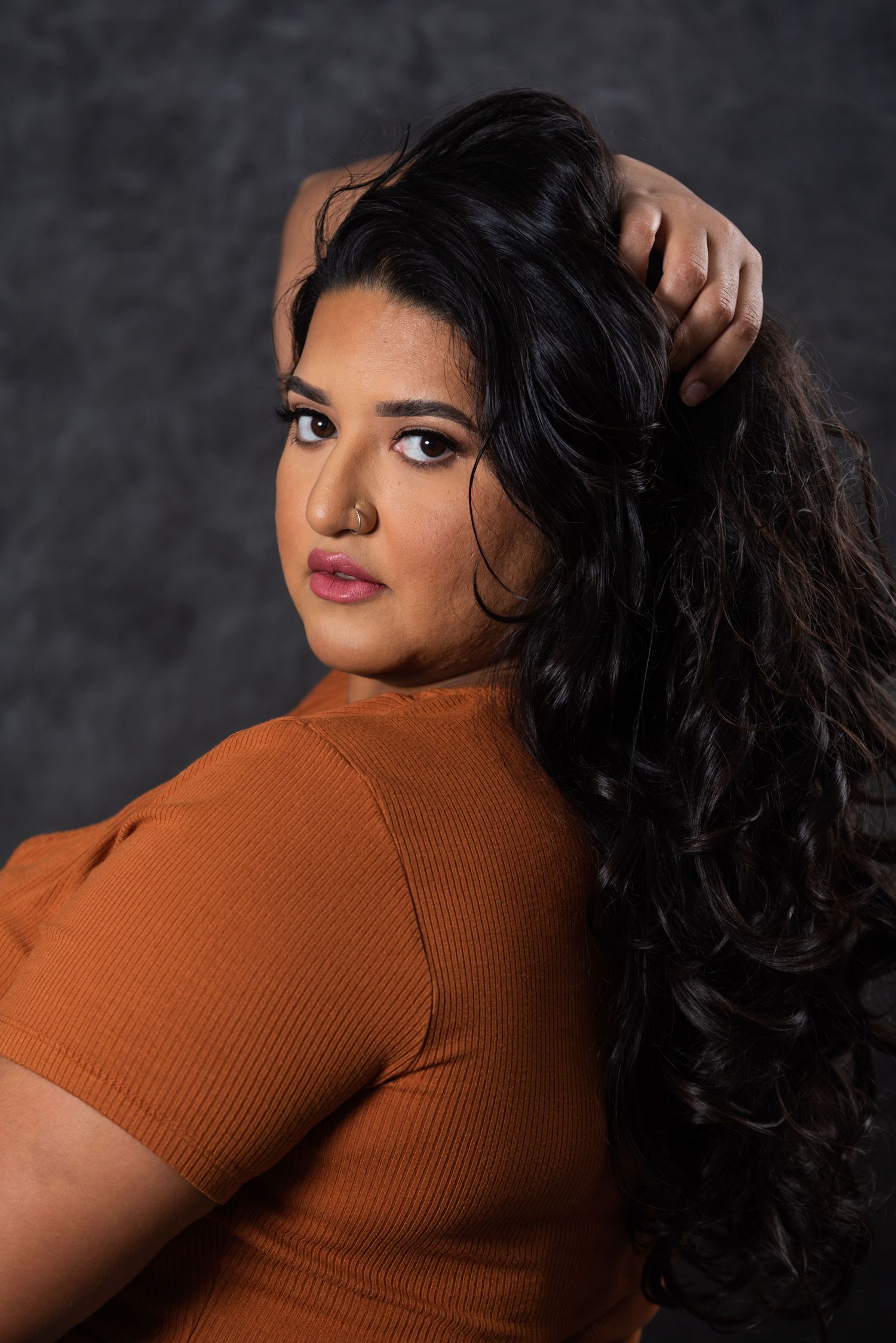 Fashion portrait in rust-colored top with long dark curls against moody grey background.