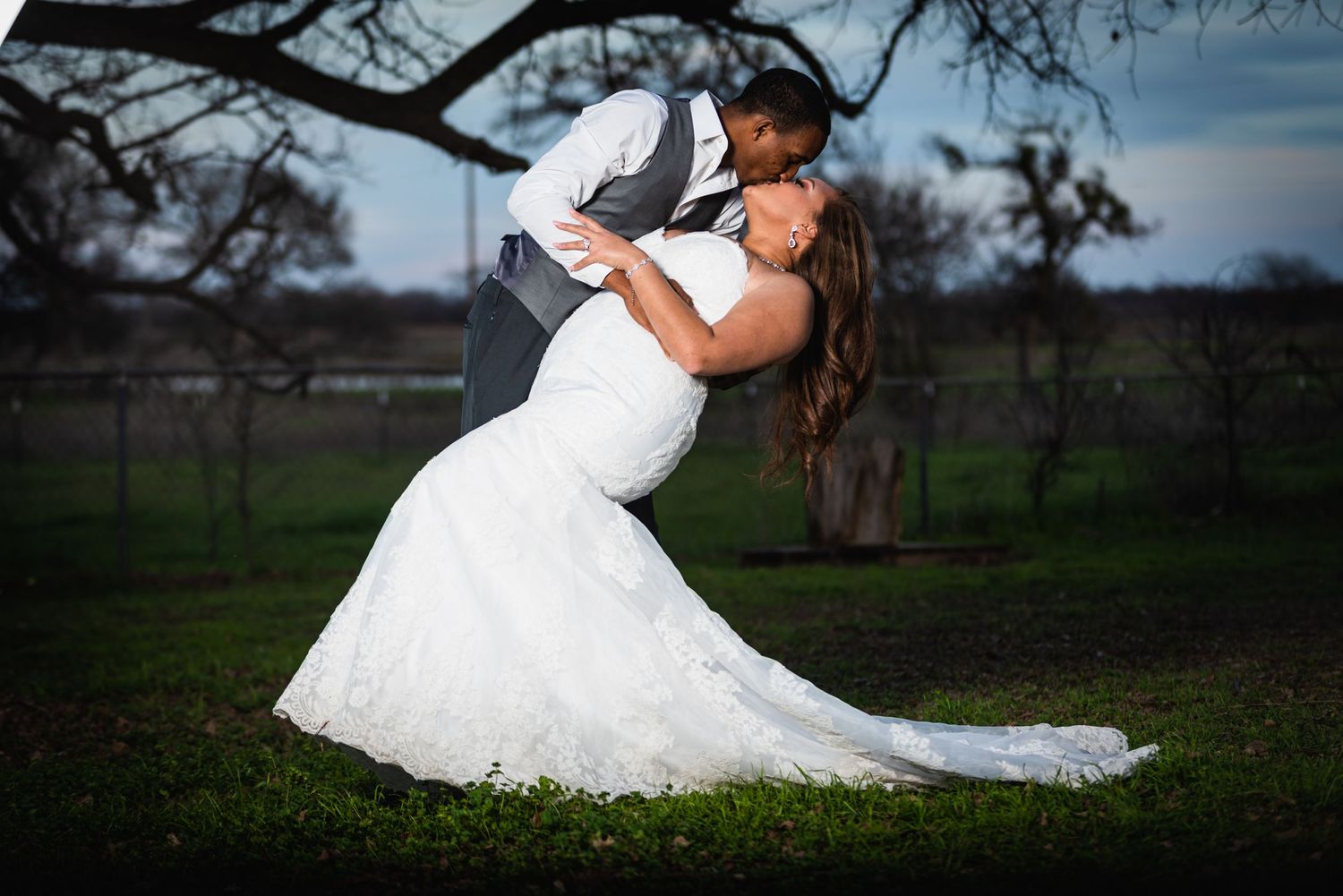 Romantic wedding photo showing dramatic dip pose with bride's white lace dress trailing against rural outdoor setting.
