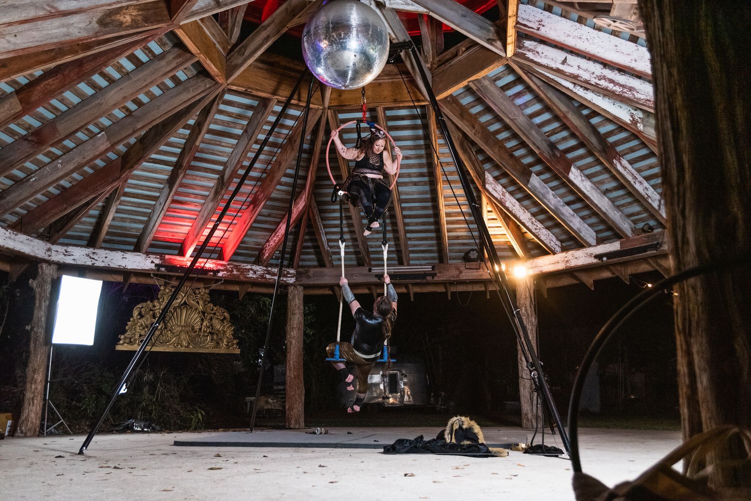 Aerial performer suspended from ceiling in dramatic lighting within wooden structure.
