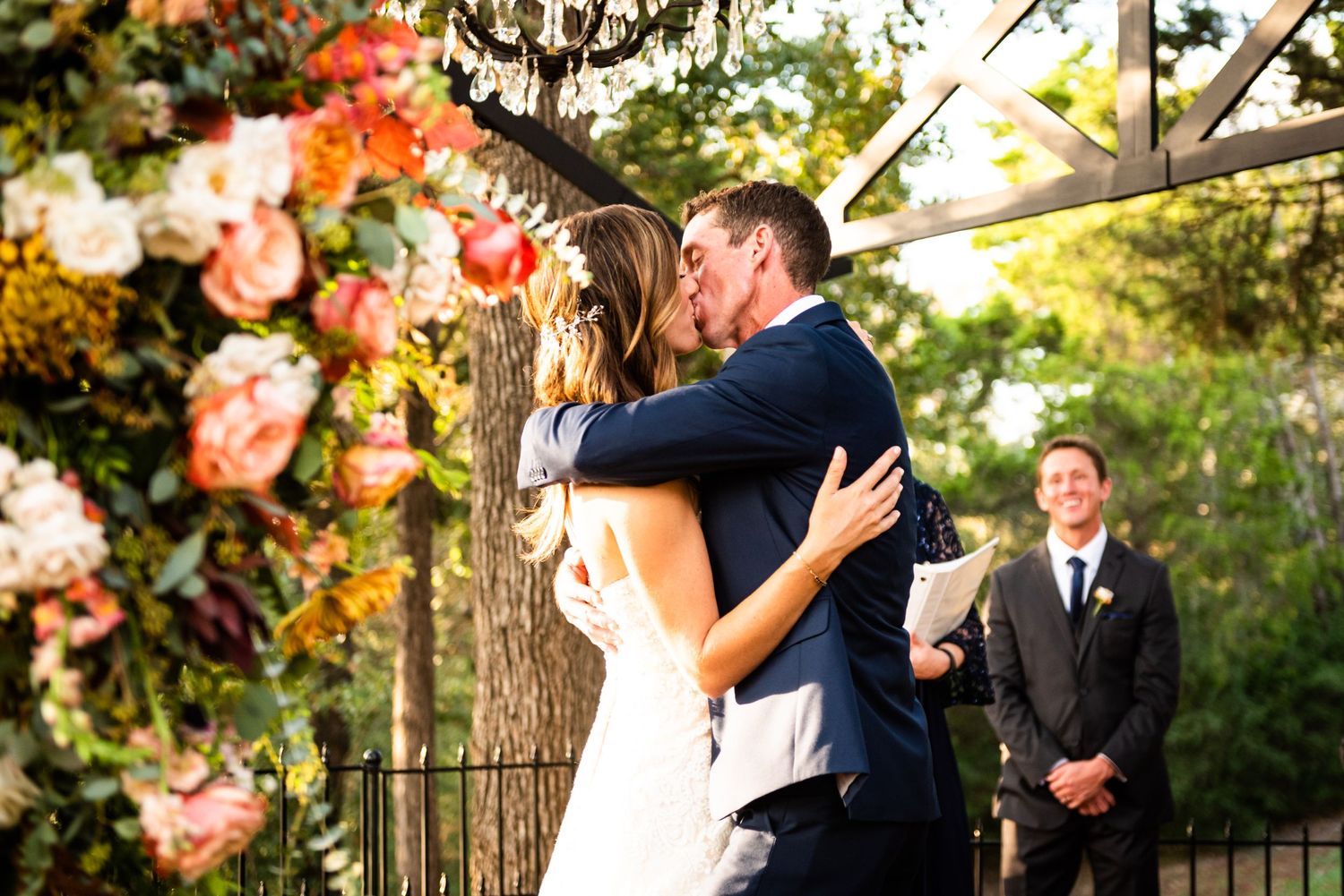 A romantic wedding kiss under a floral arch decorated with coral and pink roses during an outdoor ceremony.