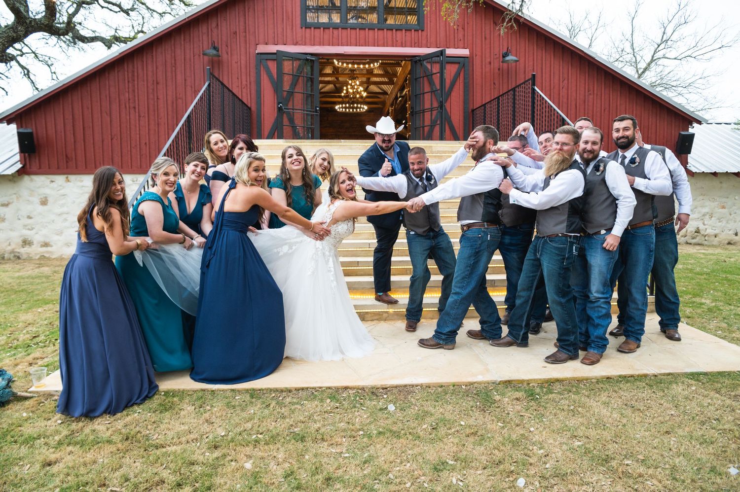 Fun wedding party photo with bridesmaids in blue and groomsmen striking playful poses outside barn.