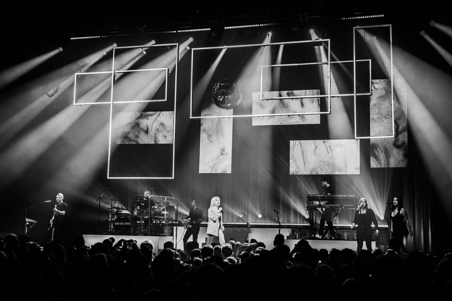 Stage performance with dramatic lighting effects and geometric patterns projected behind a live band.