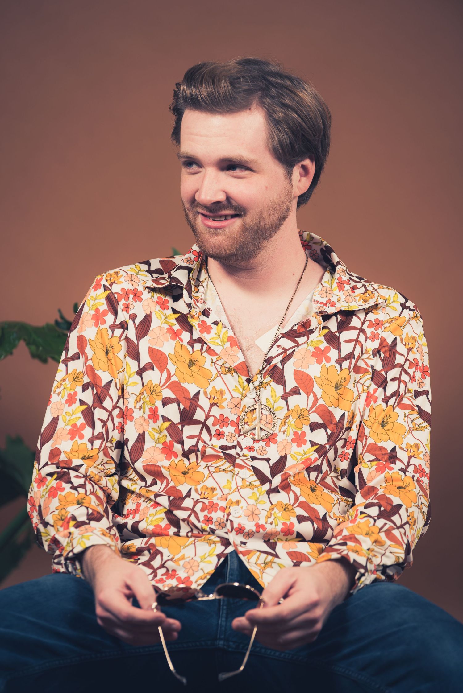 Person in colorful floral shirt sits and smiles against brown studio background.