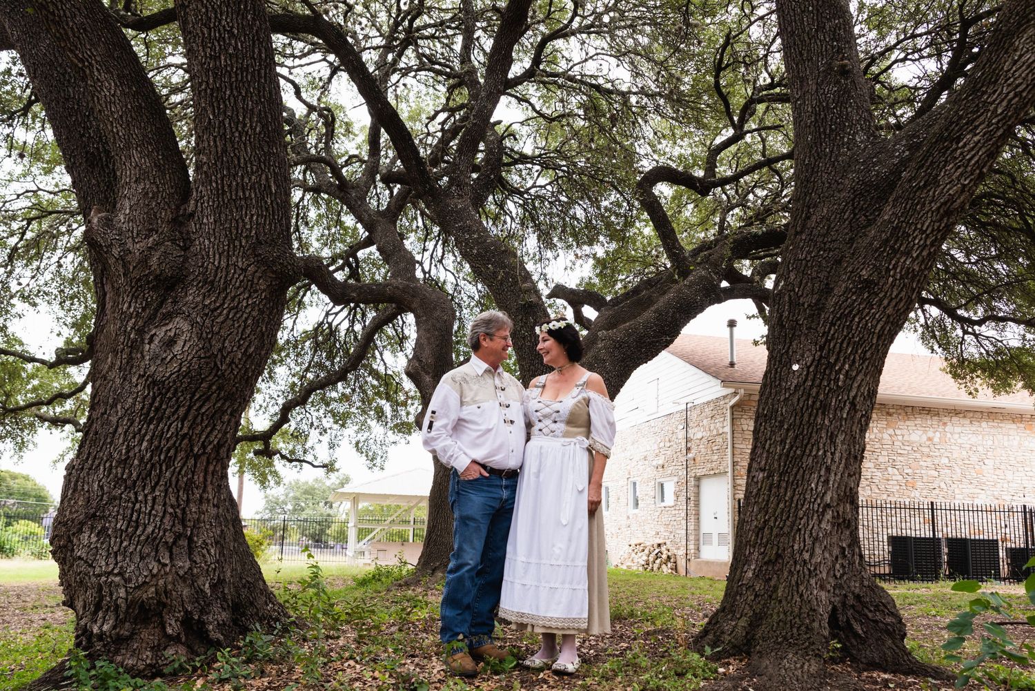 A couple poses together under majestic oak trees in rustic wedding attire.