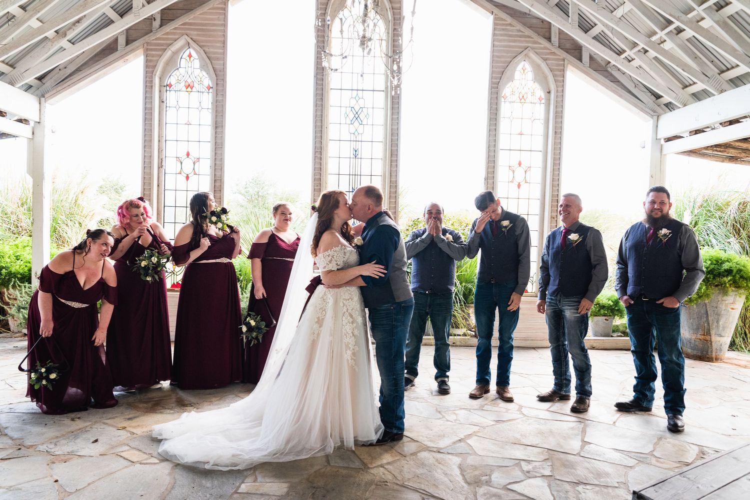 Wedding party in burgundy and blue gather in a chapel with stained glass windows.