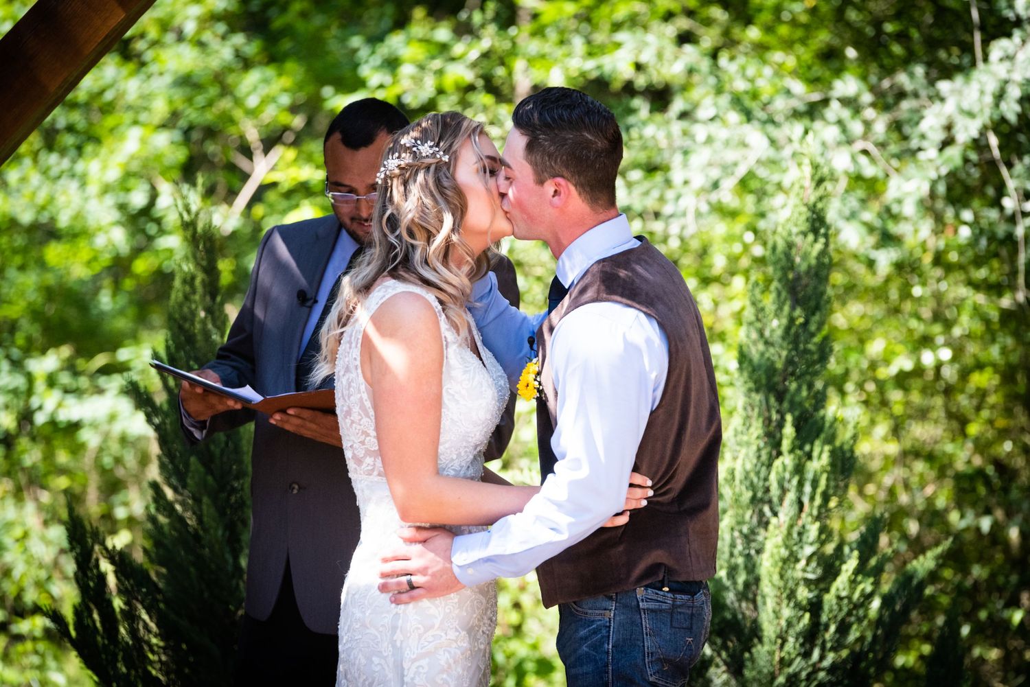 Intimate wedding ceremony kiss moment under wooden pergola surrounded by lush greenery.