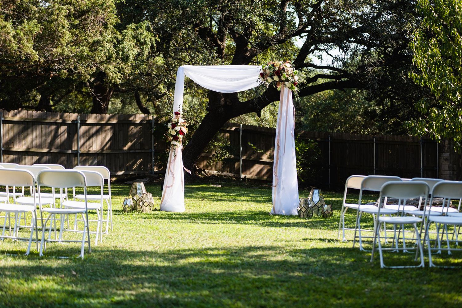 Outdoor wedding ceremony setup with white arch and folding chairs arranged on green lawn.