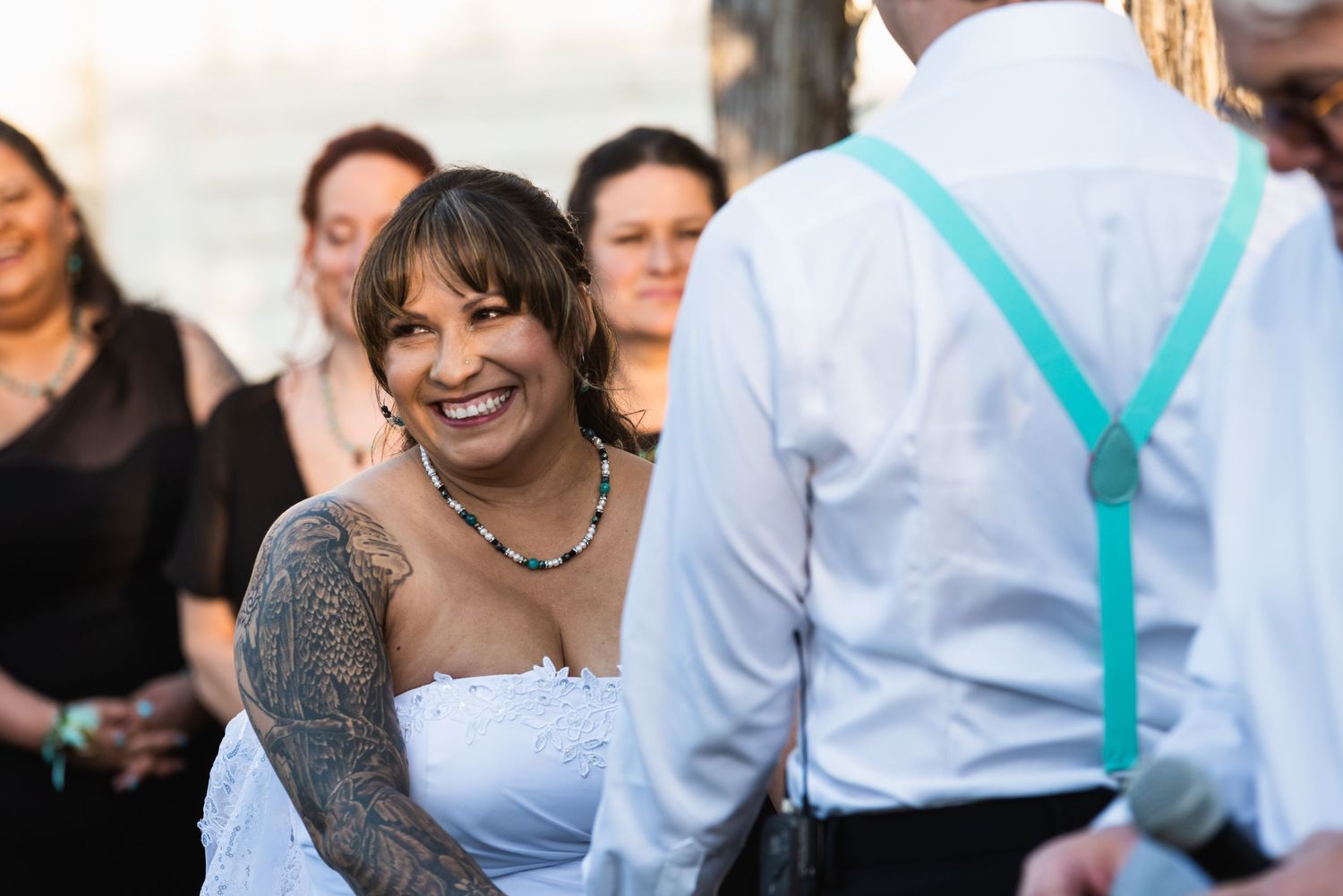 A bride in a white dress with tattoos smiles joyfully during an outdoor wedding ceremony.
